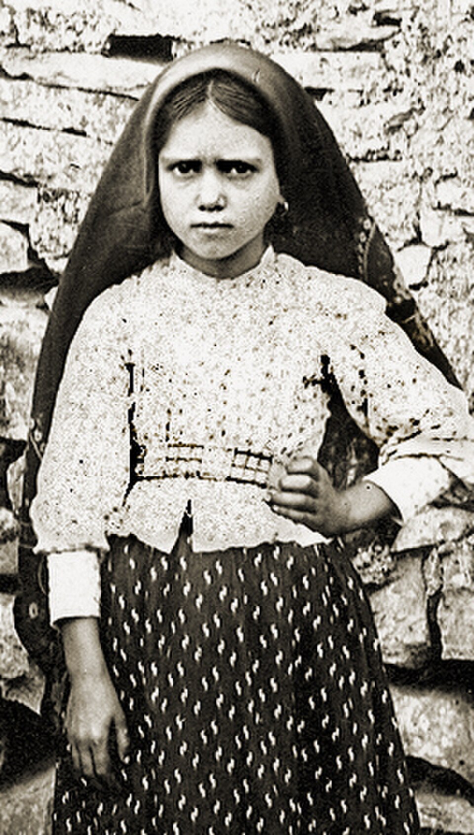 Fotografía antigua de santa Jacinta Marto niña, con pañuelo oscuro en la cabeza, delante de un muro de piedra