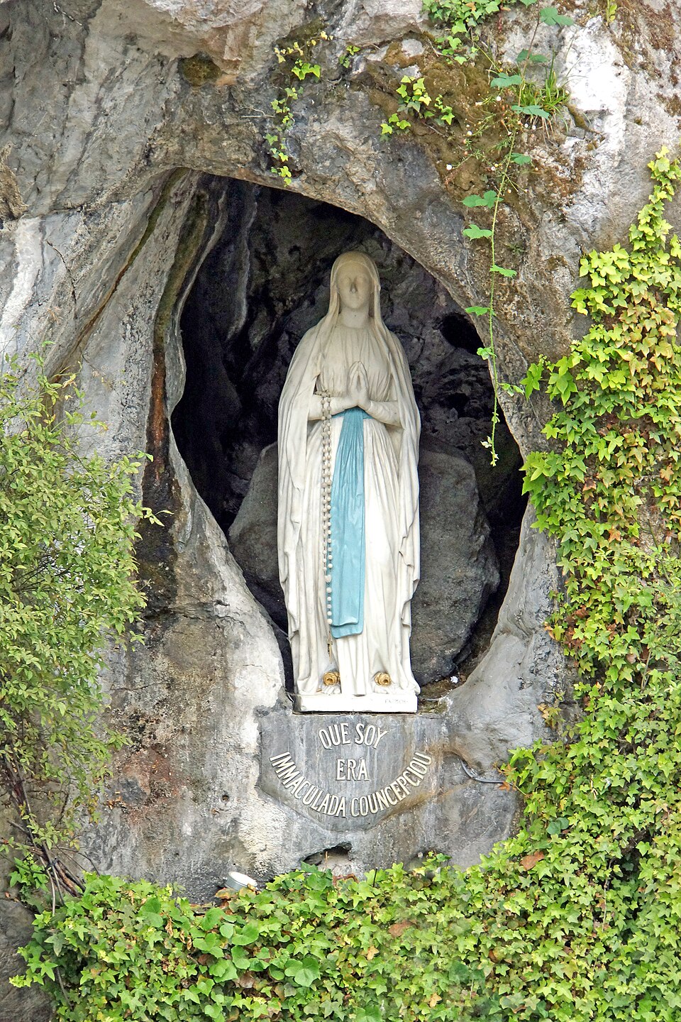 Estatua de Nuestra Señora de Lourdes en la gruta, con vestido blanco, cinta azul, manos juntas y rosario