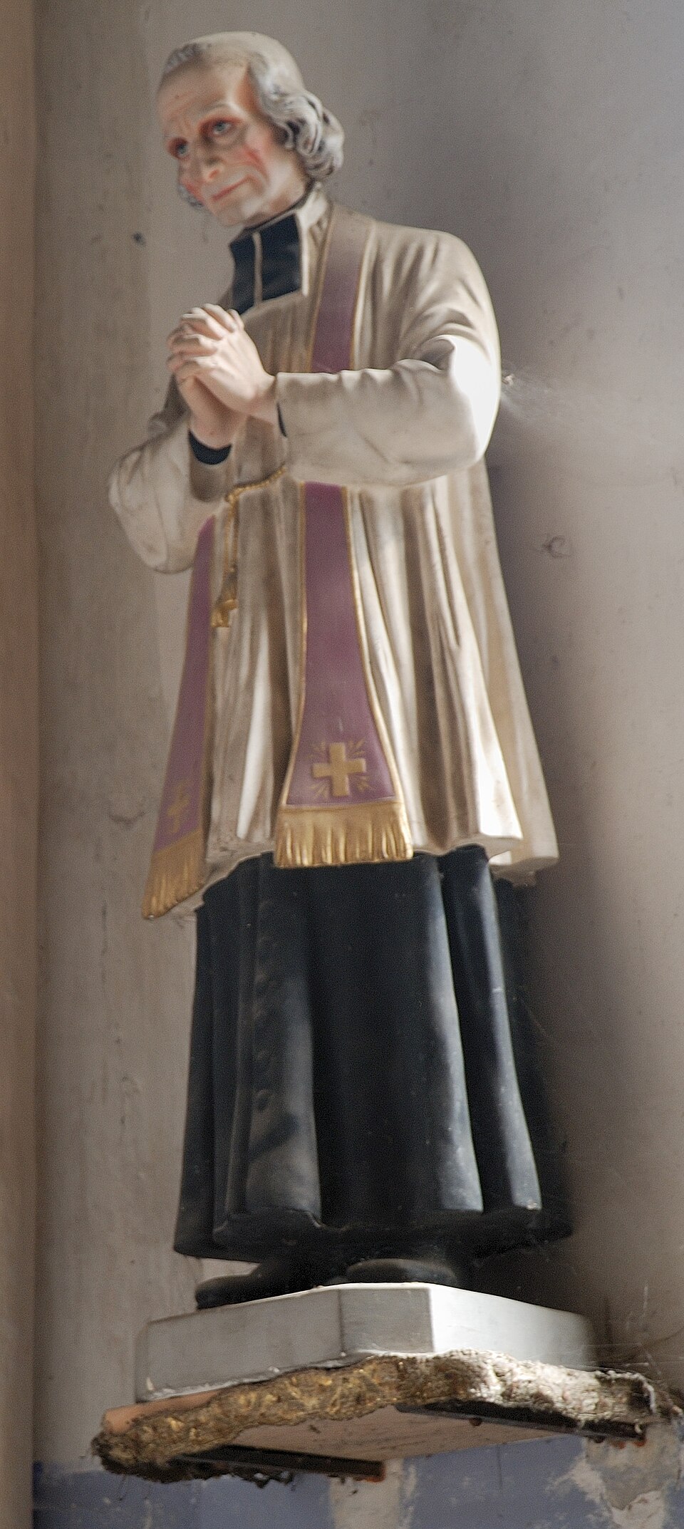 Escultura de san Juan María Vianney con sotana negra, sobrepelliz y estola morada, con las manos juntas en oración