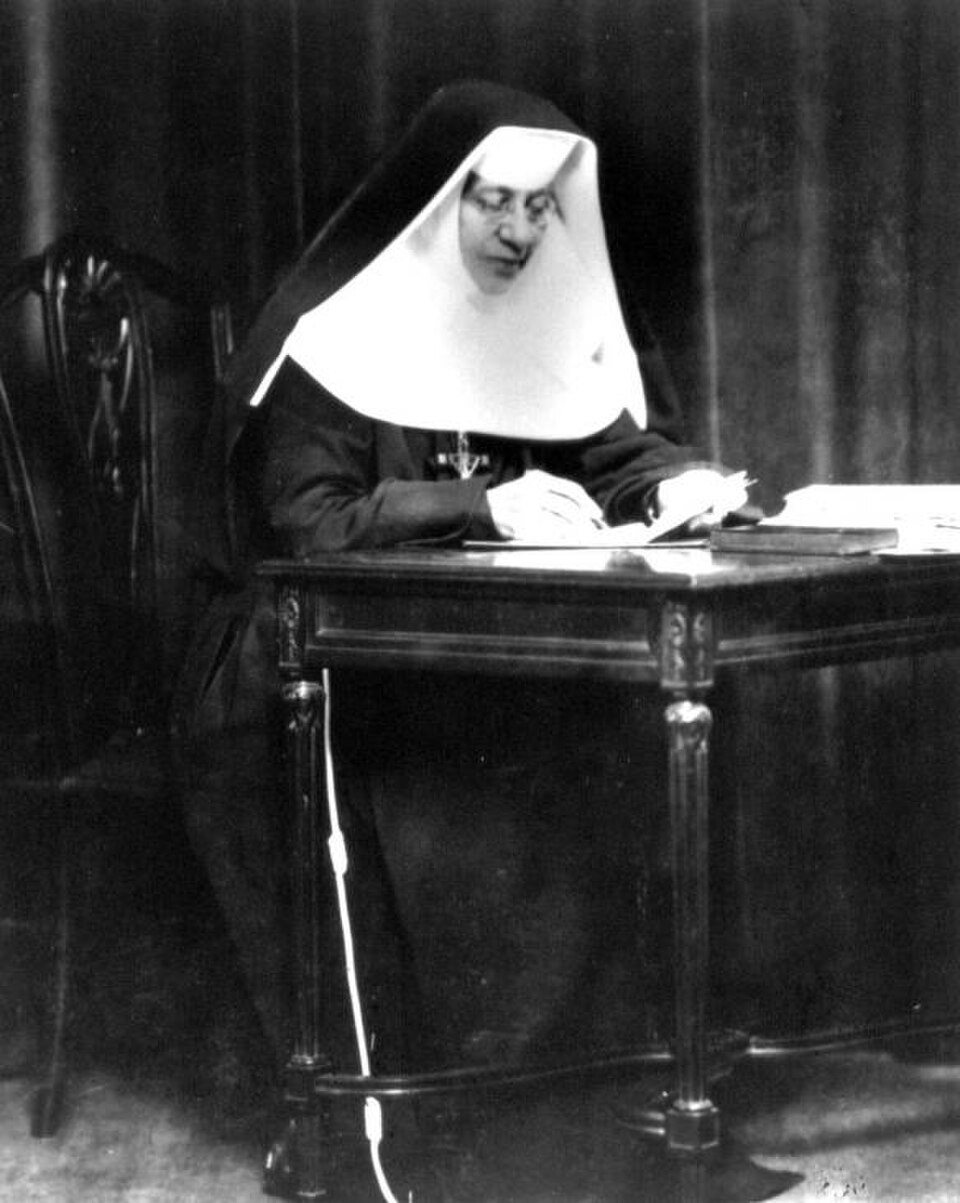 Fotografía antigua de santa Catalina Drexel con hábito y toca de religiosa, sentada leyendo ante una mesa