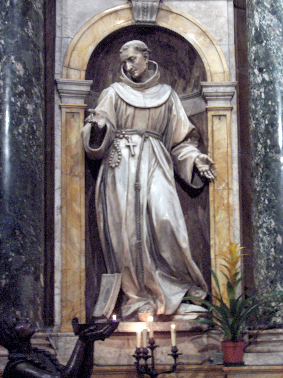 Estatua de mármol de san Bernardino de Siena con hábito franciscano y cordón, en una hornacina con velas
