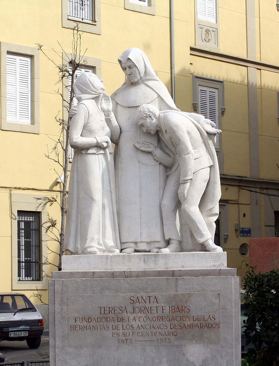 Monumento de mármol blanco a santa Teresa Jornet con hábito, acogiendo a una anciana y a un anciano