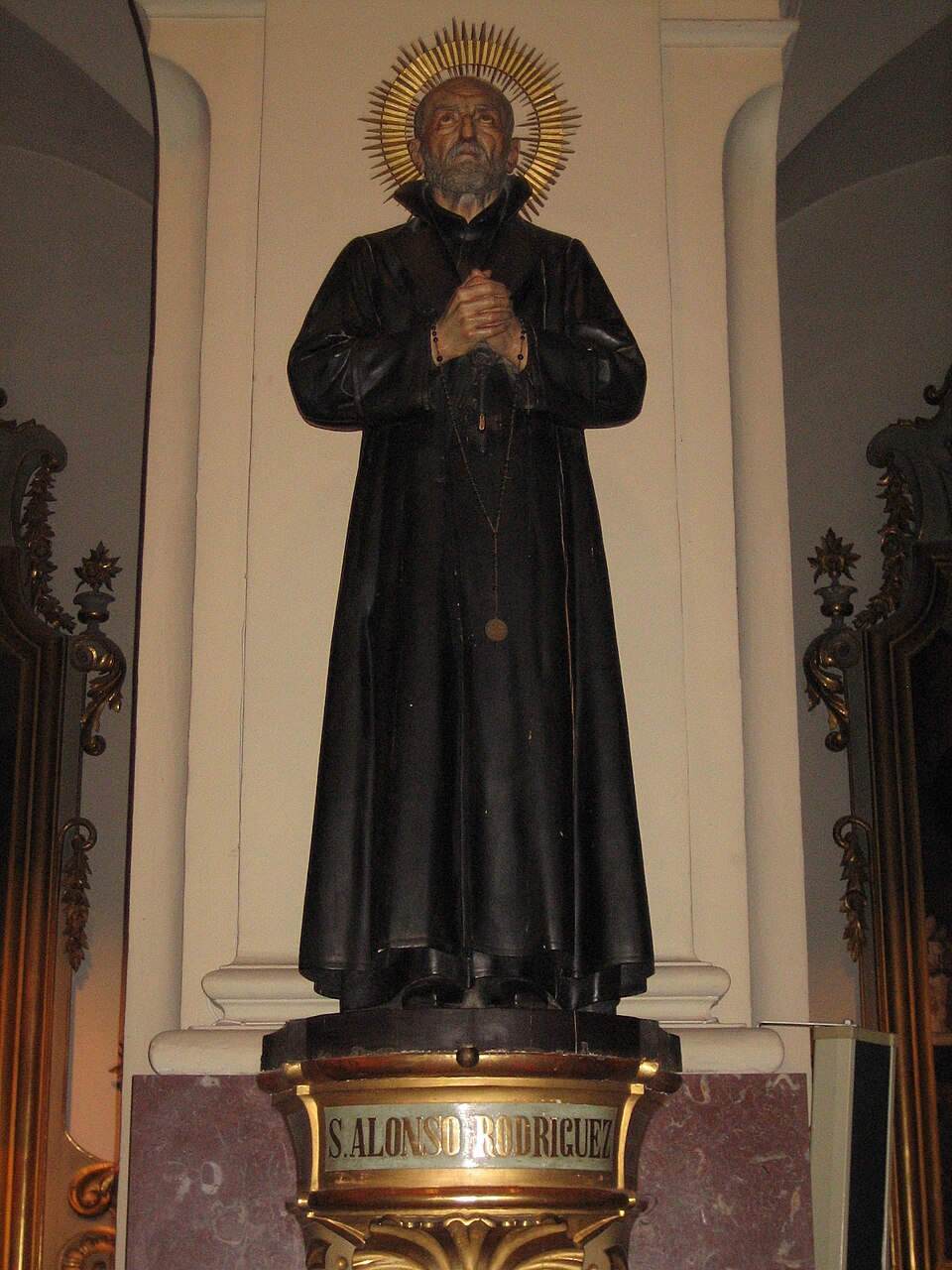 Escultura de san Alonso Rodríguez con sotana negra, aureola dorada y rosario, con las manos juntas en oración